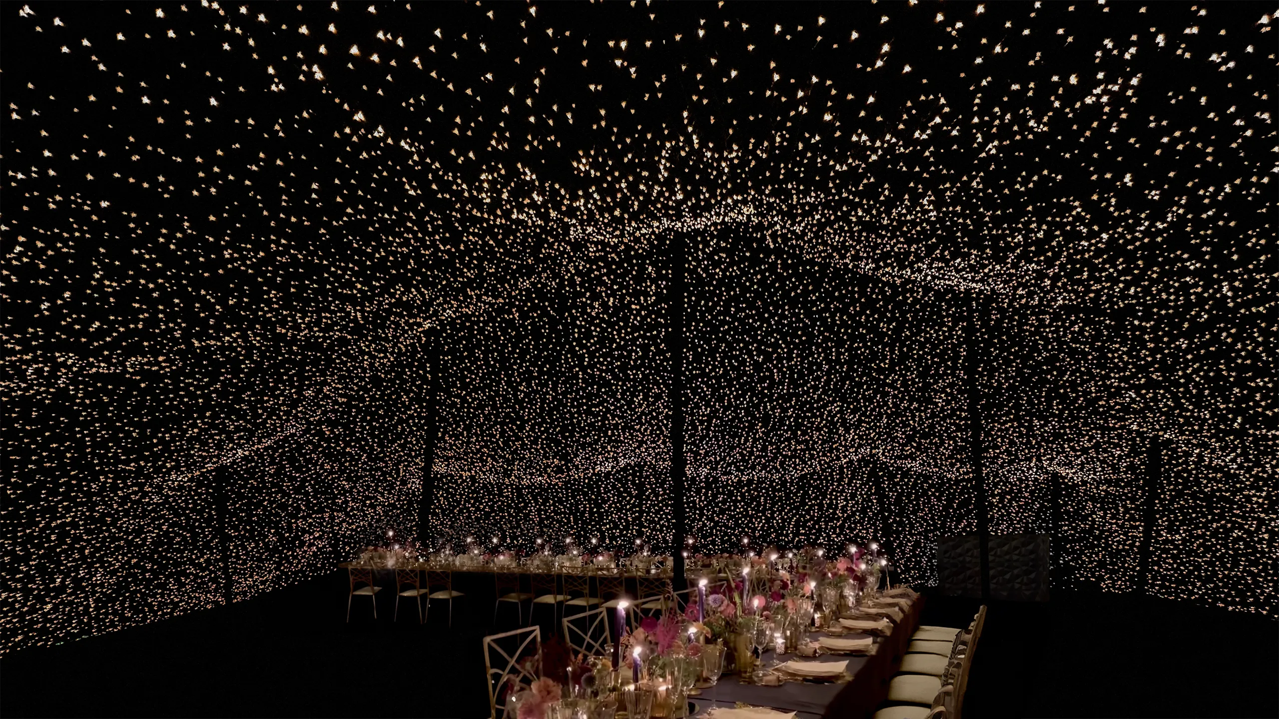 Interior de Carpa Beduina negra cerrada totalmente hasta el suelo con millones de luces en el interior a modo de estrellas drante la celebracion de una fiesta con mesas decoradas con flores rosas, velas, vajilla elegante, cuberteria dorada y sillas doradas a juego con la iluminacion.