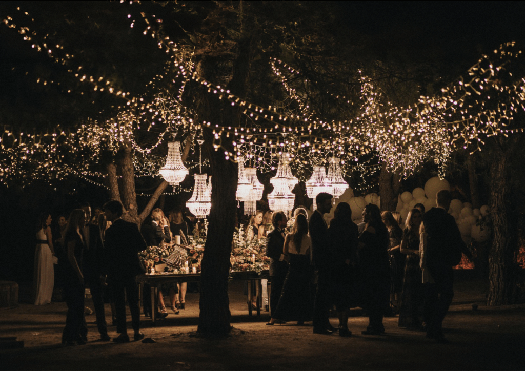 Una elegante recepción nocturna al aire libre bajo árboles decorados con hileras de luces brillantes y chandeliers de cristal colgantes, con invitados vestidos de etiqueta conversando alrededor de una mesa decorada.
