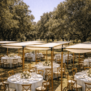Varias mesas redondas con manteles blancos y centros de flores, dispuestas bajo módulos individuales de pérgolas rústicas de madera en un bosque o jardín con árboles.