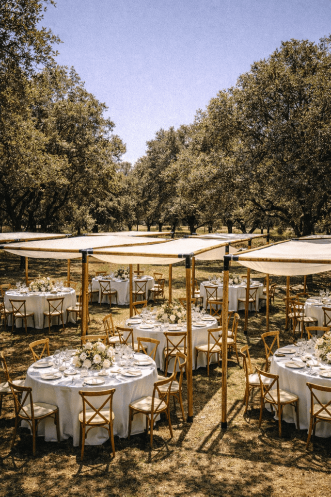Varias mesas redondas con manteles blancos y centros de flores, dispuestas bajo módulos individuales de pérgolas rústicas de madera en un bosque o jardín con árboles.