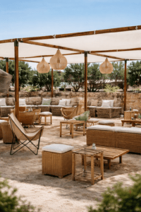 Salón exterior bajo pérgola de madera con sofás de bambú, cojines verdes y blancos, pufs cuadrados y lámparas de flecos sobre suelo de grava.