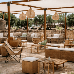 Salón exterior bajo pérgola de madera con sofás de bambú, cojines verdes y blancos, pufs cuadrados y lámparas de flecos sobre suelo de grava.