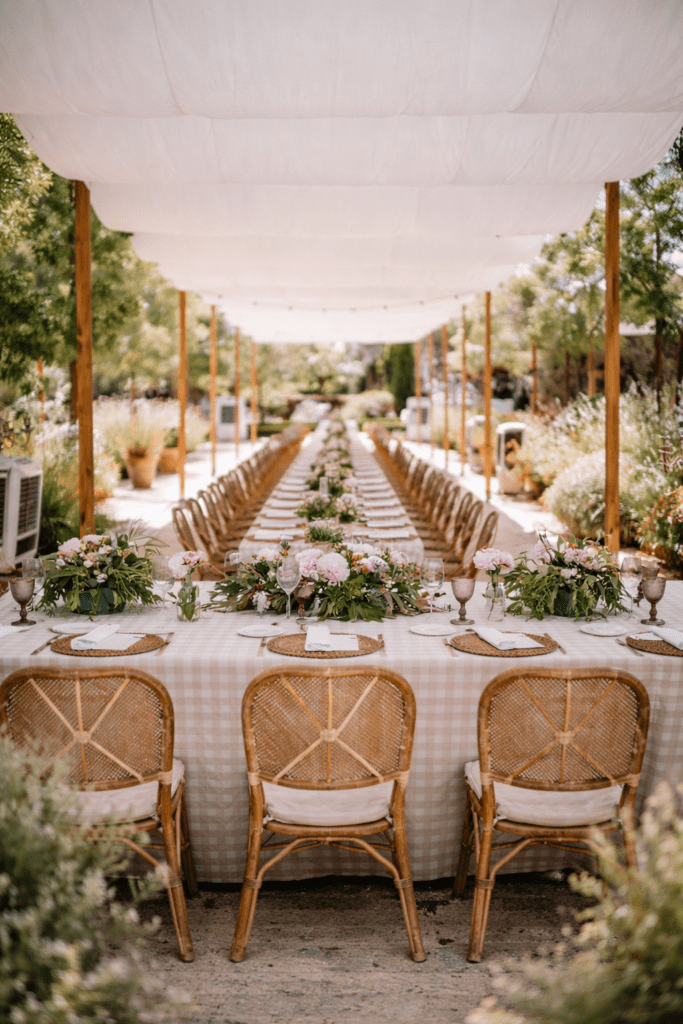 Mesa imperial para eventos decorada con mantel de cuadros vichy beige, centros de flores rosas y sillas de rejilla bajo una pérgola con lonas blancas drapeadas.