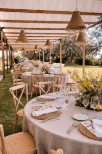 Montaje de banquete exterior con mesas redondas, mantelería de lino, sillas Crossback y lámparas de esparto bajo una pérgola de madera con toldos vela blancos.