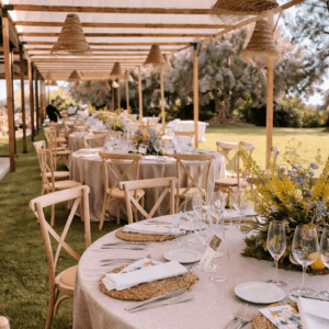 Montaje de banquete exterior con mesas redondas, mantelería de lino, sillas Crossback y lámparas de esparto bajo una pérgola de madera con toldos vela blancos.