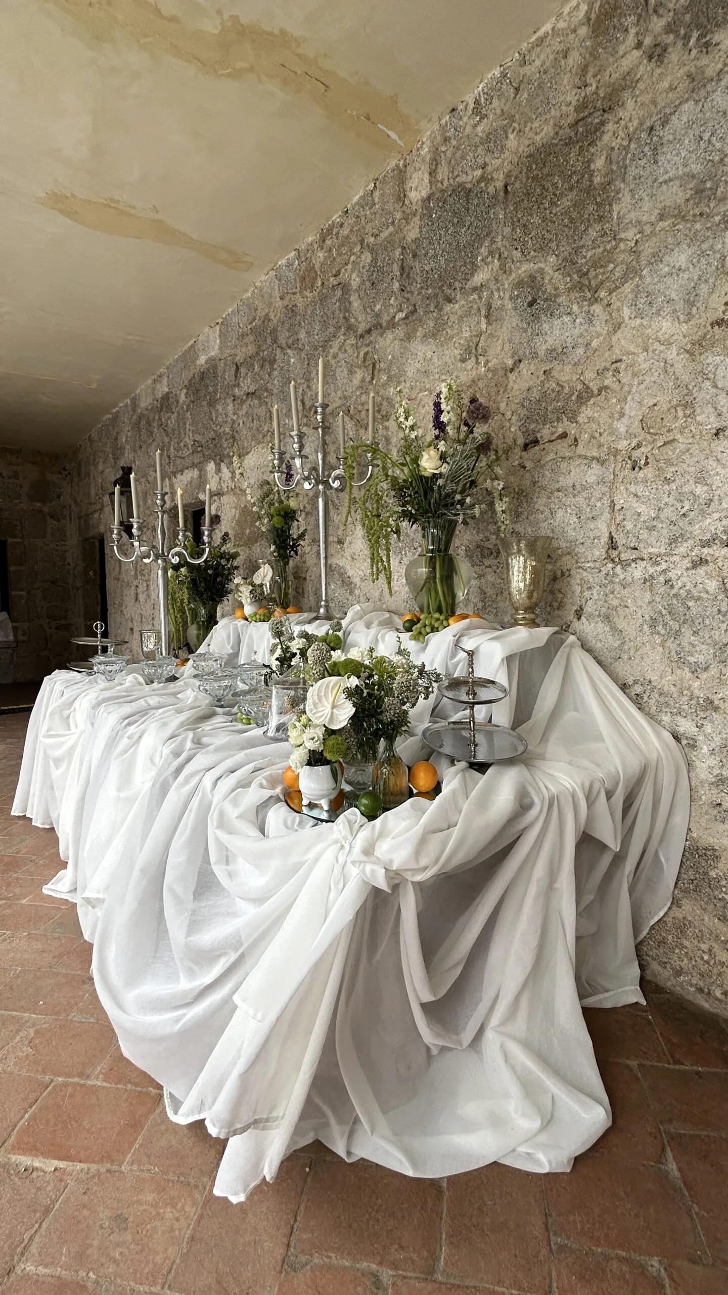 Primer plano de mesa con mantelería blanca drapeada de forma vaporosa, decorada con candelabros de plata y flores blancas contra una pared de piedra.