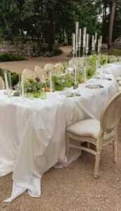 Primer plano de mesa de banquete decorada con mantelería blanca drapeada, candelabros de plata, arreglos florales y frutas cítricas sobre suelo de barro.