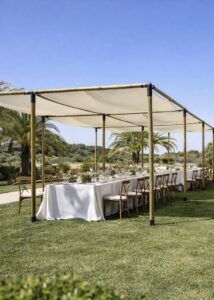 Estructura de pérgola de madera con toldo de lona blanca tensada cubriendo una mesa imperial con mantelería blanca y sillas Crossback en un jardín con palmeras.
