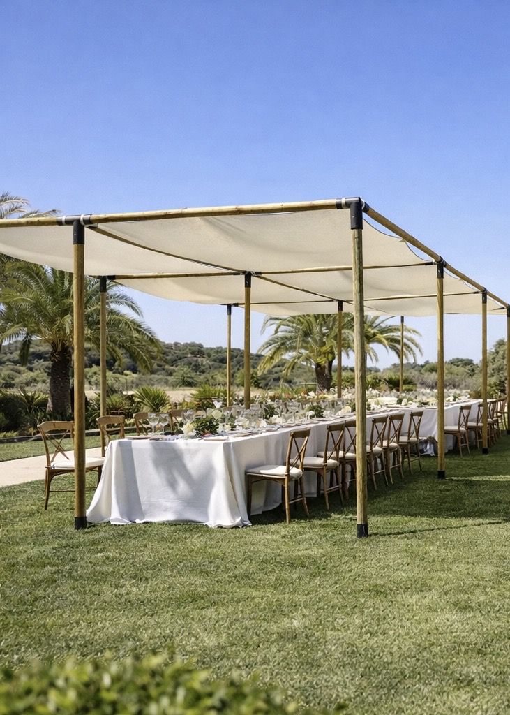 Estructura de pérgola de madera con toldo de lona blanca tensada cubriendo una mesa imperial con mantelería blanca y sillas Crossback en un jardín con palmeras.