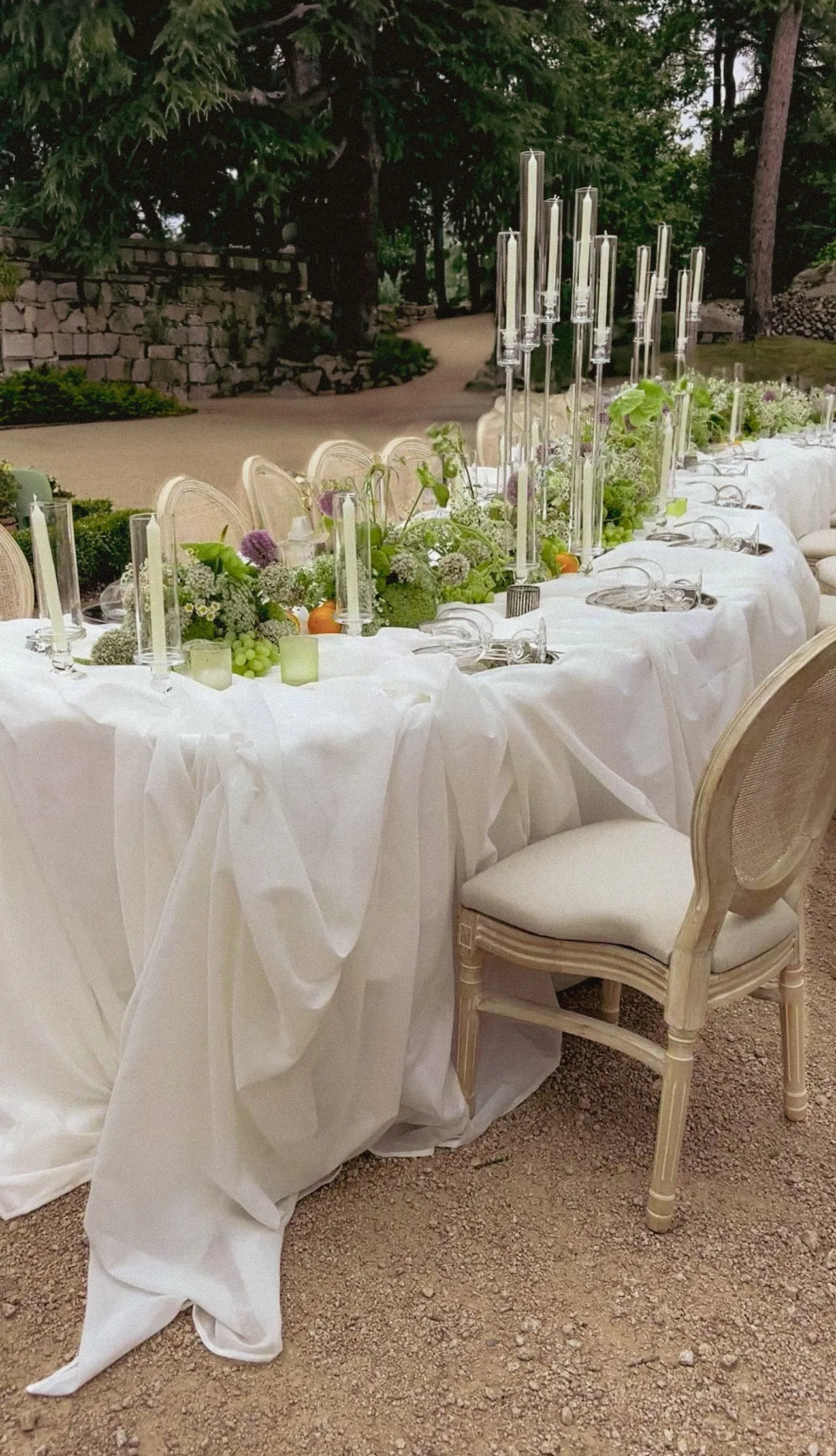 Primer plano de mesa de banquete decorada con mantelería blanca drapeada, candelabros de plata, arreglos florales y frutas cítricas sobre suelo de barro.